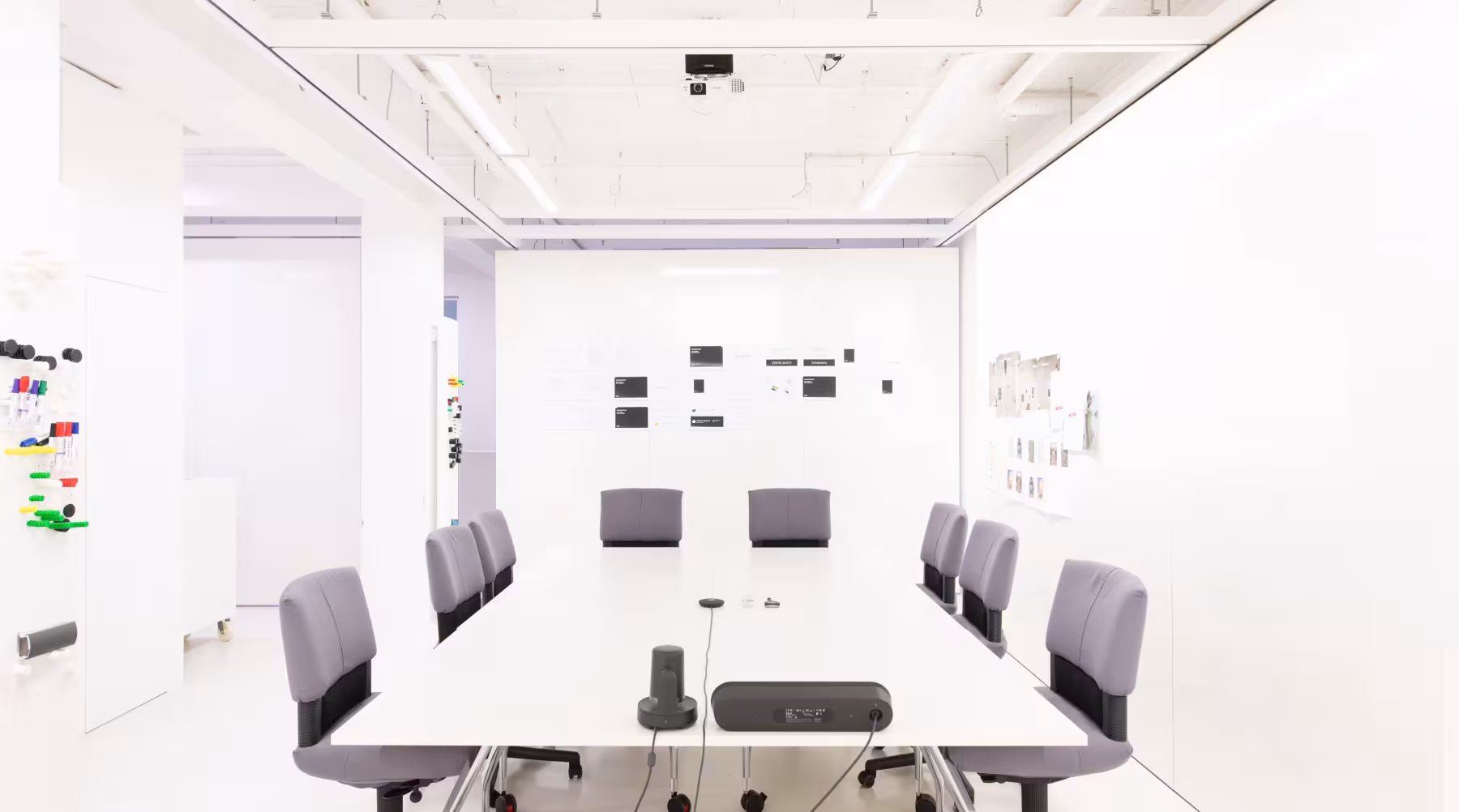 Heller, moderner Besprechungsraum mit wei&szlig;en W&auml;nden, Decke und Boden. In der Mitte steht ein gro&szlig;er, wei&szlig;er Konferenztisch mit acht gepolsterten, grauen Drehst&uuml;hlen. Auf dem Tisch befindet sich Konferenztechnik. An den W&auml;nden sind verschiedene Notizen, Skizzen und Plakate befestigt. Die Beleuchtung ist klar und freundlich, an der Decke h&auml;ngt ein Beamer. Der Raum wirkt kreativ, aufger&auml;umt und einladend.
