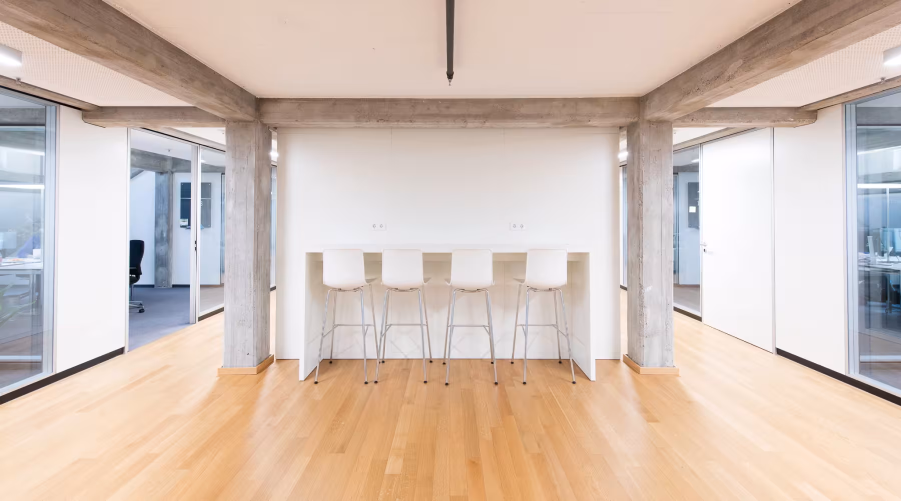 Heller, moderner B&uuml;roraum mit Holzfu&szlig;boden und Sichtbeton-Elementen. Im Zentrum stehen f&uuml;nf wei&szlig;e Barhocker an einem hohen wei&szlig;en Tisch, der an einer wei&szlig;en Wand befestigt ist. Links und rechts sind Glasw&auml;nde und T&uuml;ren zu angrenzenden B&uuml;ror&auml;umen sichtbar, in denen Schreibtische und B&uuml;rost&uuml;hle stehen. Der Raum wirkt offen, minimalistisch und freundlich, mit klaren Linien und viel Tageslicht.