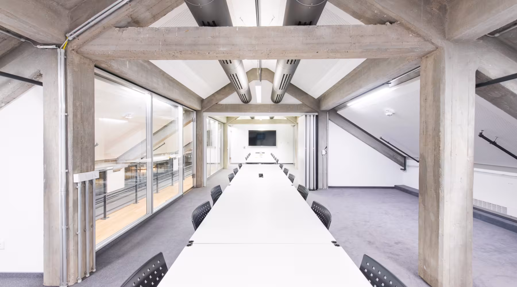 Heller, moderner Konferenzraum mit schr&auml;gen Decken und sichtbaren Betontr&auml;gern. In der Mitte steht ein langer, wei&szlig;er Konferenztisch mit vielen schwarzen St&uuml;hlen. Am Ende des Raumes h&auml;ngt ein gro&szlig;er Flachbildschirm an der Wand. Links verl&auml;uft eine Glaswand mit Blick auf einen Flur oder eine Galerie, rechts ist die Wand schr&auml;g und wei&szlig;. Der Raum wirkt gro&szlig;z&uuml;gig, aufger&auml;umt und ist technisch ausgestattet.
