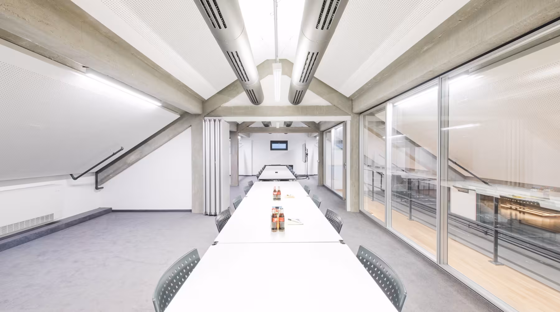 Heller, moderner Konferenzraum mit schr&auml;gen wei&szlig;en Decken und sichtbaren Betontr&auml;gern. In der Mitte steht ein langer, wei&szlig;er Konferenztisch mit grauen St&uuml;hlen. Auf dem Tisch stehen mehrere Flaschen und Gl&auml;ser, sowie Notizbl&ouml;cke. Rechts verl&auml;uft eine gro&szlig;e Glaswand mit Blick auf einen Flur oder eine Galerie, am Ende des Raumes h&auml;ngt ein Flachbildschirm an der Wand. Der Raum wirkt gro&szlig;z&uuml;gig, aufger&auml;umt und einladend.