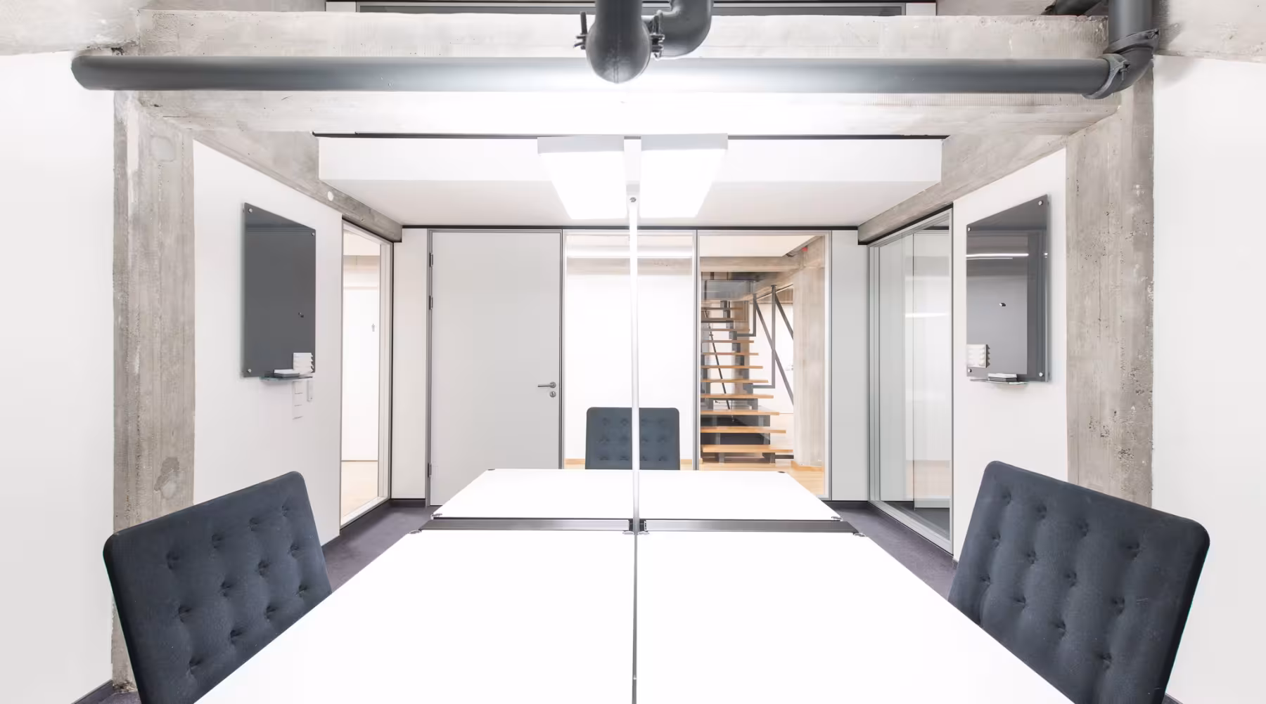 Heller, moderner B&uuml;roraum mit wei&szlig;en W&auml;nden, sichtbaren Betontr&auml;gern und offenliegenden Rohren an der Decke. In der Mitte stehen zwei gro&szlig;e, wei&szlig;e Schreibtische mit vier gepolsterten, dunklen B&uuml;rost&uuml;hlen. Im Hintergrund f&uuml;hren offene Holztreppen nach oben, seitlich sind Glast&uuml;ren und kleine Wandtafeln zu sehen. Der Raum wirkt aufger&auml;umt, minimalistisch und freundlich.