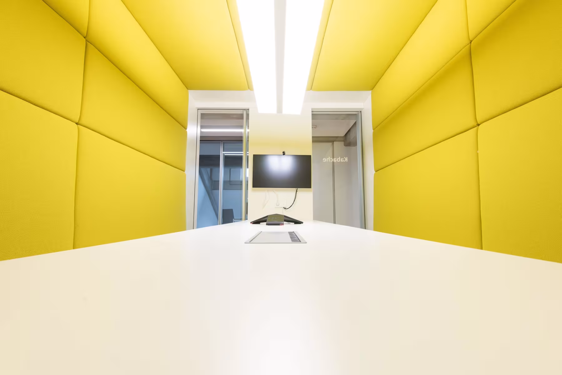 Kleiner, moderner Besprechungsraum mit auff&auml;lligen, gelben, gepolsterten W&auml;nden. In der Mitte steht ein langer, wei&szlig;er Tisch. An der Stirnseite h&auml;ngt ein Flachbildschirm mit Videokonferenzsystem an der Wand. Durch Glast&uuml;ren sind angrenzende R&auml;ume sichtbar. Der Raum wirkt hell, freundlich und schallged&auml;mmt.