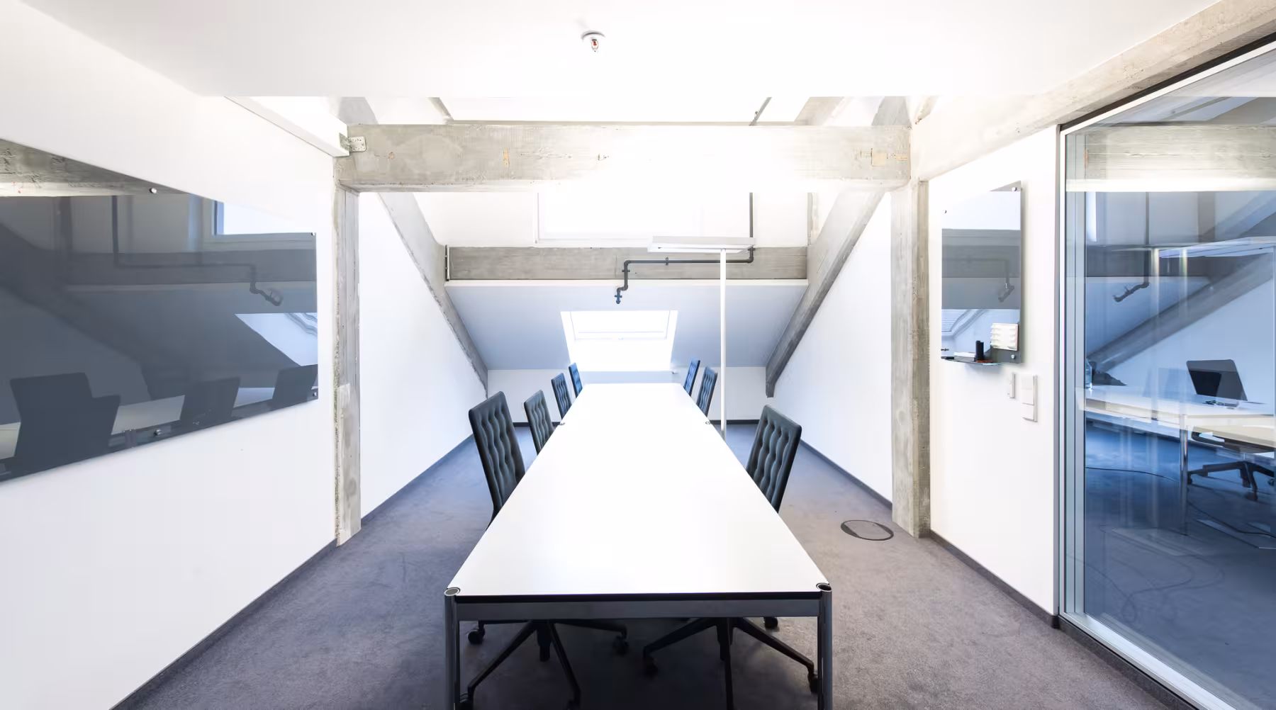 Meetingraum im Dachgeschoss mit langem wei&szlig;en Konferenztisch, acht gepolsterten St&uuml;hlen, Sichtbetontr&auml;gern, Dachfenstern und einer Glaswand zum Flur.