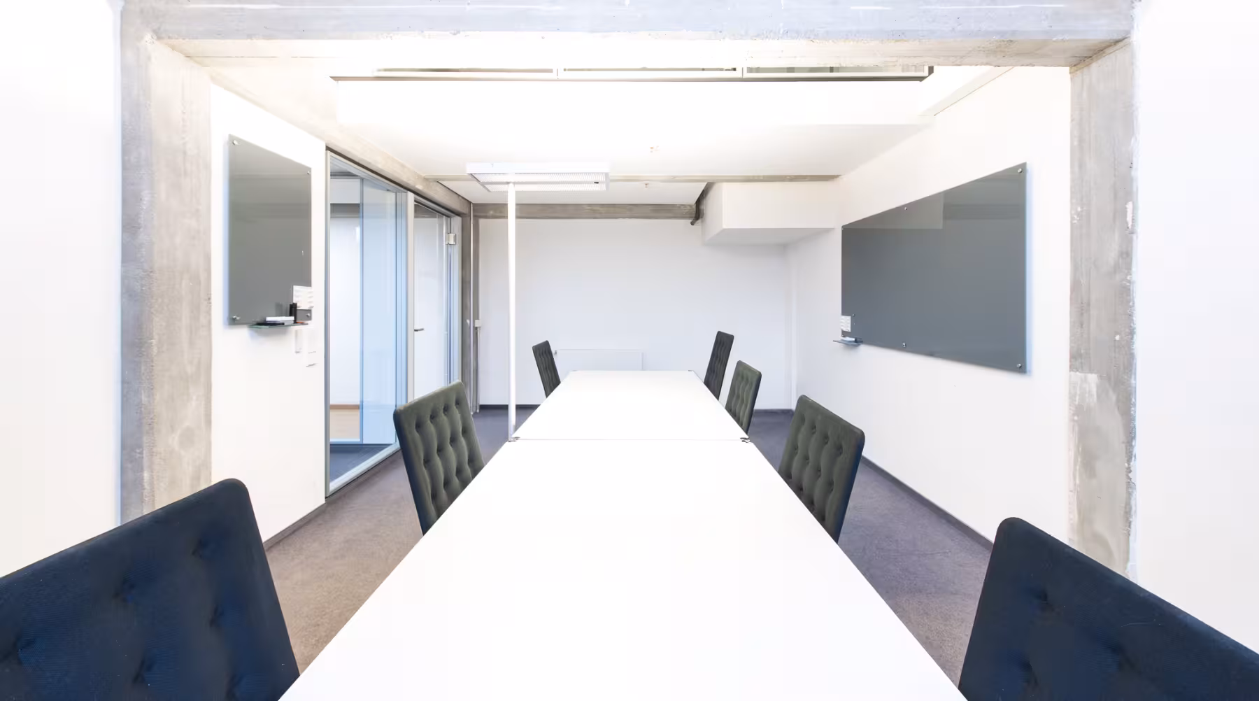 Konferenzraum mit langem wei&szlig;em Tisch, gepolsterten St&uuml;hlen in Blau und Grau, zwei Whiteboards und Sichtbetonelementen an der Decke.