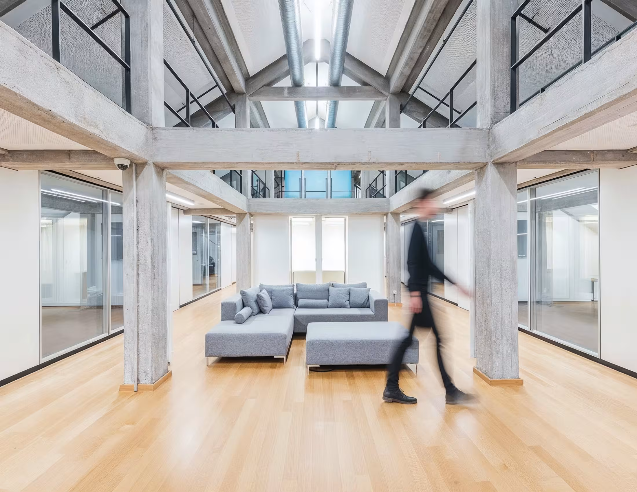 Moderner, heller Eingangsbereich eines B&uuml;ros mit Holzfu&szlig;boden und Sichtbeton-Tr&auml;gern. Im Zentrum steht eine gro&szlig;e, graue Eckcouch mit mehreren Kissen und einem passenden Hocker. Im Hintergrund sind gro&szlig;e Fenster und Glast&uuml;ren zu sehen, seitlich befinden sich Glasw&auml;nde zu angrenzenden B&uuml;ros. Eine Person in dunkler Kleidung geht unscharf durchs Bild, was dem Raum eine lebendige Atmosph&auml;re verleiht. Oben ist eine offene Galerie mit Metallgel&auml;nder sichtbar.