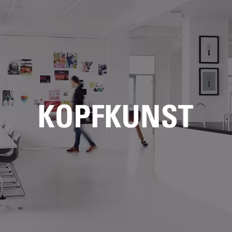 Heller, moderner B&uuml;roraum mit wei&szlig;em Interieur, gro&szlig;em Tisch und mehreren St&uuml;hlen. Im Hintergrund sind verschiedene bunte Bilder und Plakate an der Wand angebracht. Rechts befindet sich eine offene K&uuml;chenzeile und eine Treppe mit wei&szlig;en Gel&auml;ndern. Mehrere Personen bewegen sich durch den Raum. Mittig im Bild steht gro&szlig; und wei&szlig; der Schriftzug &bdquo;KOPFKUNST&ldquo;.